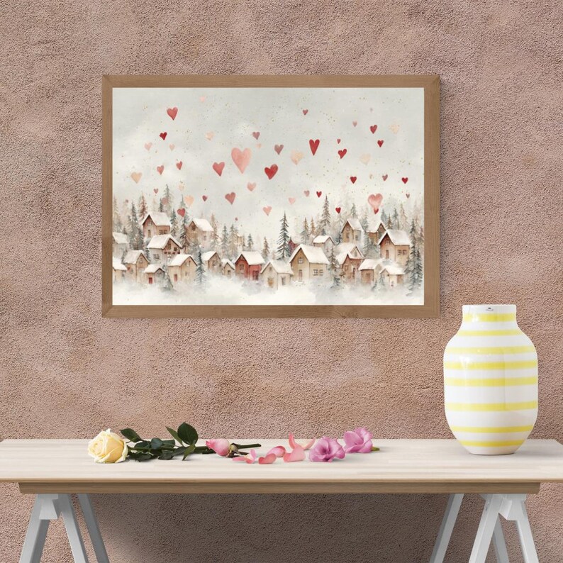 May include: Watercolor painting of a winter village scene with red hearts floating in the sky. The artwork features small houses nestled among evergreen trees, framed in a light brown wooden frame. A yellow and white striped vase sits on a wooden table.