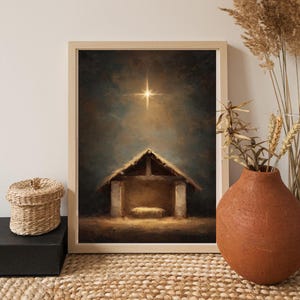 Op de afbeelding: Een ingelijste prent toont een kerststal met een ster boven een rustieke stal. Het kunstwerk heeft een warm kleurenpalet, met de nadruk op het licht van de ster en de eenvoudige schuilplaats. De prent wordt getoond op een geweven oppervlak, met een decoratieve mand en vaas in de buurt.