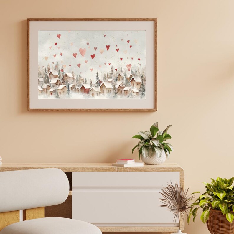 May include: Watercolor print of a winter village scene with snow-covered rooftops and evergreen trees. Red and pink hearts float in the sky above the village. The artwork is framed in a light brown wooden frame.