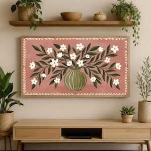 May include: A framed floral art print featuring a vase with white flowers and green leaves against a dusty rose background. The vase is olive green, and the frame is a light wood color. The artwork is displayed on a wooden console table.