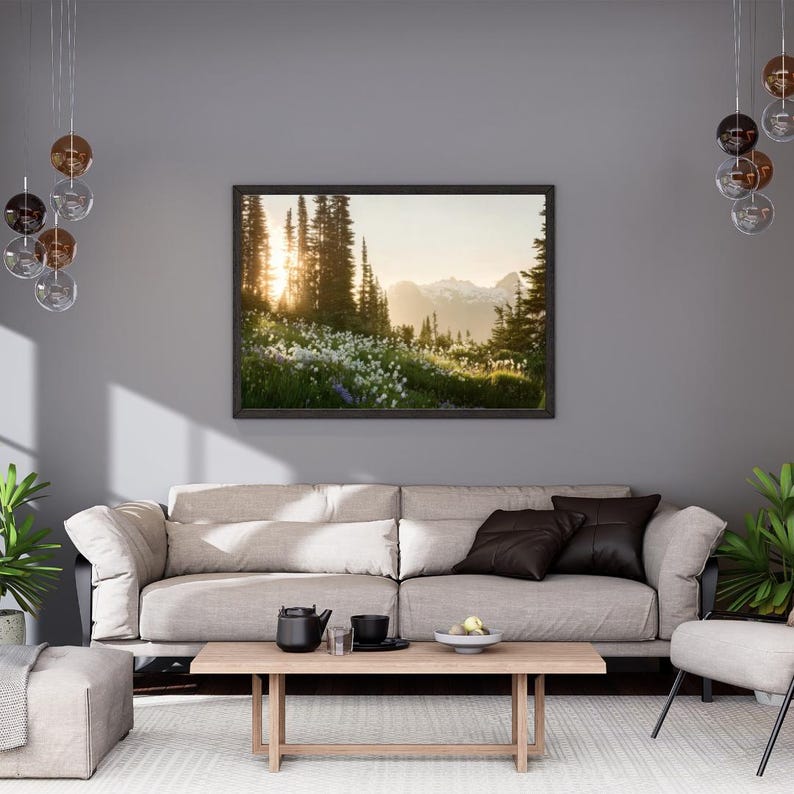 May include: A framed landscape photograph of a mountain scene with tall trees and a bright sunset. The image is displayed above a light-colored sofa with dark pillows, a wooden coffee table, and decorative lighting.