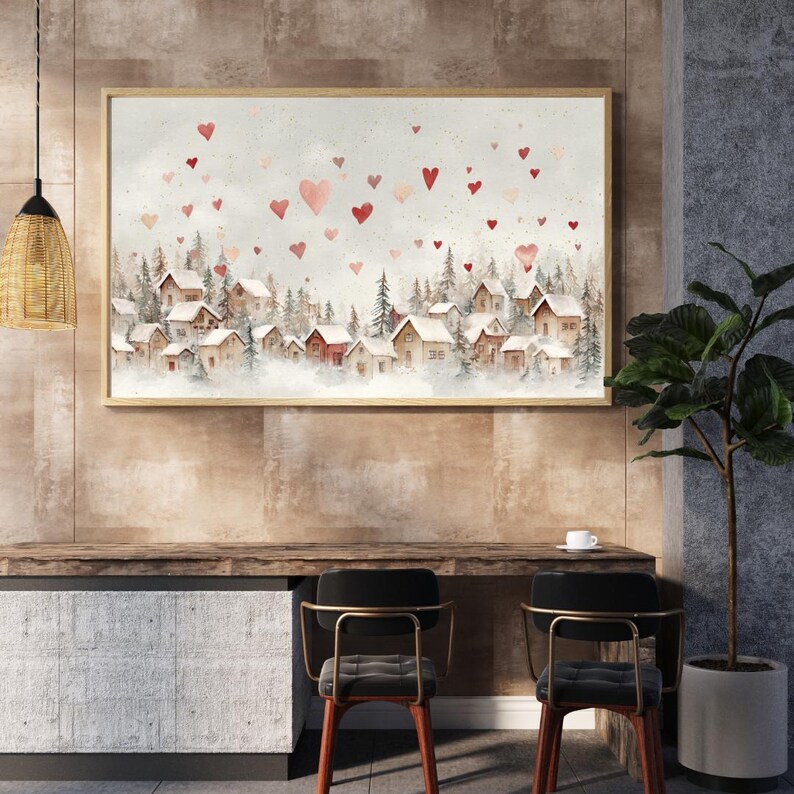 May include: A framed watercolor painting depicts a snow-covered village with red-roofed houses and pine trees. Red and pink hearts float in the sky above the village, creating a romantic and whimsical scene. The artwork is displayed above a wooden desk.