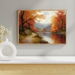 May include: A framed landscape painting showing an autumnal scene with a river reflecting the orange and red leaves of the trees. A path follows the riverbank, leading into the distance, with mountains in the background. The artwork is in a wooden frame.