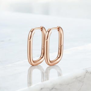 Könnte beinhalten: Ein Paar roségoldene Creolen. Die Ohrringe haben eine rechteckige Form mit abgerundeten Kanten. Die Ohrringe werden auf einer weißen Marmoroberfläche präsentiert.