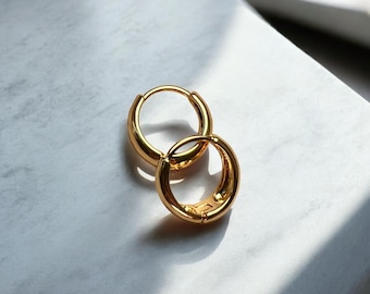 May include: A pair of gold-colored hoop earrings. The earrings are round with a thick band and a secure clasp. The earrings are displayed on a white and gray surface.