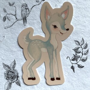 May include: A sticker featuring a light blue deer with white spots and a light brown background. The deer is standing on all four legs and has its head turned to the side. The deer has a light brown nose and a light brown mouth.