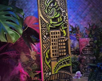 May include: A rectangular wooden tiki carving featuring a green and brown monster destroying a building. The carving has intricate details and is surrounded by tropical plants and flowers. The carving is approximately 24 inches tall.