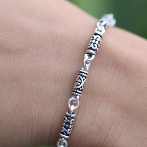 Könnte beinhalten: Ein silbernes Armband mit detaillierten, aufwendigen Mustern. Das Armband verfügt über dekorative zylindrische Perlen, die durch kleinere Glieder verbunden sind. Das Design umfasst geprägte Muster und einen klassischen, eleganten Stil. Das Armband wird vor einem verschwommenen Hintergrund gezeigt.