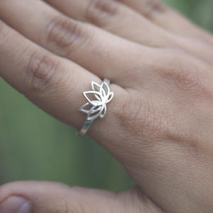 May include: A silver ring featuring a delicate lotus flower design. The ring is worn on a finger, showcasing the intricate openwork petals. The ring's band is thin and simple, complementing the floral centerpiece. The ring is a piece of jewelry.