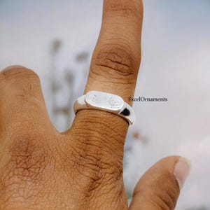 Puede incluir: Un anillo de plata con una cara rectangular con un diseño de sol. El anillo se lleva en un dedo. El texto "ExcelOrnaments" es visible en el lado derecho de la imagen.
