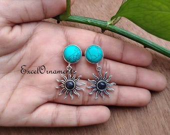 May include: A pair of earrings featuring turquoise-colored round stones at the top and sunburst pendants with a dark center. The earrings are silver-toned and are displayed against a wooden surface.