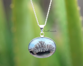 May include: A silver pendant necklace with an oval-shaped pendant. The pendant features a landscape design with a tree-like pattern in shades of black and white against a light blue background. The necklace is suspended from a delicate silver chain.