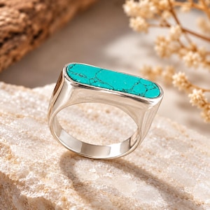 May include: A silver ring featuring a horizontal, rectangular inlay of bright turquoise stone with dark veining. The metal band has a smooth, polished finish and a flat top surface that holds the elongated, vibrant blue-green gemstone securely.