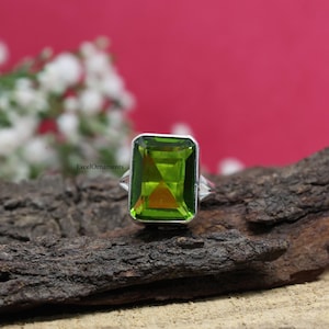 May include: A silver ring featuring a large, rectangular, bright green gemstone. The gemstone has a faceted cut, and the ring is set against a dark, textured wooden surface with a blurred pink background.