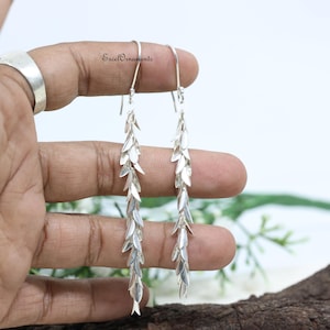 May include: Silver drop earrings featuring a cascading leaf design. The earrings are long and slender, with multiple small, leaf-shaped charms hanging from a hook. The earrings are displayed against a light background.