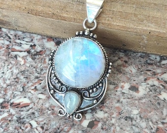 Op de afbeelding: Zilveren hanger met een grote, ronde, iriserende maansteen en een kleinere, druppelvormige maansteen. De hanger heeft een sierlijke zilveren zetting met een oogje voor een ketting. De maansteen vertoont een blauwe en witte kleur.