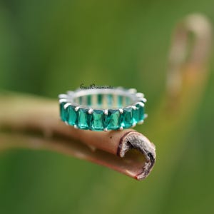 Peut inclure: Une bague en argent ornée de pierres précieuses vertes taille émeraude, serties en continu. La bague est présentée sur une tige brune, sur un fond vert flou. C'est un bijou.