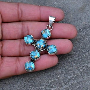 May include: A silver pendant in the shape of a cross, featuring six square turquoise stones with gold flecks. The pendant has a silver bail for a chain. The turquoise stones are set in silver bezels.