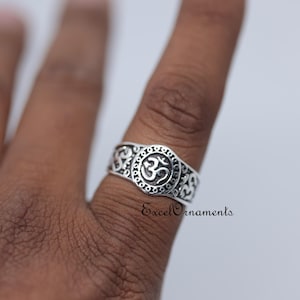 May include: A silver ring with an Om symbol in the center, surrounded by a circular design and ornate patterns. The ring is on a finger, with the text "ExcelOrnaments" visible.