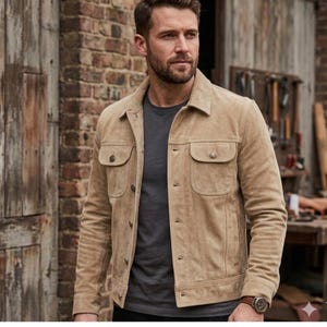 May include: A tan suede jacket with two chest pockets and two lower pockets. The jacket has a button-up front and a collared neckline. The sleeves are long and buttoned at the cuffs. The jacket is worn over a gray t-shirt.