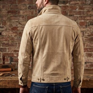 May include: A tan suede jacket featuring a structured collar, long sleeves with buttoned cuffs, and a button-tab waist adjustment. The back panel displays vertical seam detailing and a small rectangular brand label near the hem.