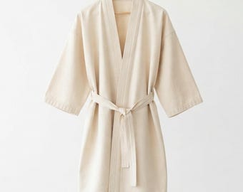 May include: A solid cream-coloured robe with wide, three-quarter length sleeves and a matching fabric tie belt at the waist. The garment features a V-neckline with a structured collar and is displayed hanging on a wooden hanger against a plain background.