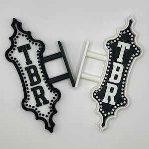 May include: Two decorative signs with the letters TBR. One sign is black with white lettering and the other is white with black lettering. Both signs have a decorative border and a stand.