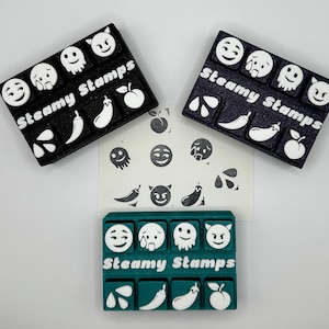 May include: Three black stamp sets with white emoji and food designs, including a ghost, aubergine, and peach. The text "Steamy Stamps" is printed on each set. A sheet of stamped images is in the centre, and a teal stamp set is at the bottom.