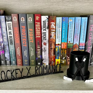 May include: A bookshelf displays a collection of books with colorful spines. A black hockey-themed pen holder with crossed hockey sticks and a "HOCKEY X ROMANCE" sign are in front of the books.