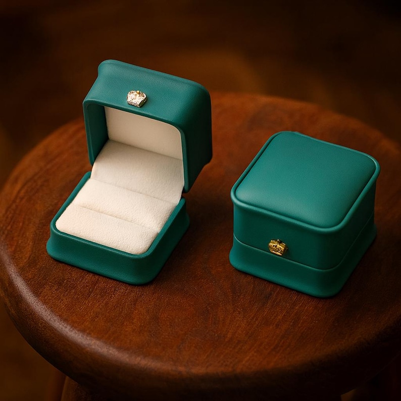 May include: Two teal-colored ring boxes, one open and one closed, sit on a wooden surface. The open box reveals a cream-colored interior with ring slots. Both boxes feature a small gold-colored crown emblem on the front.