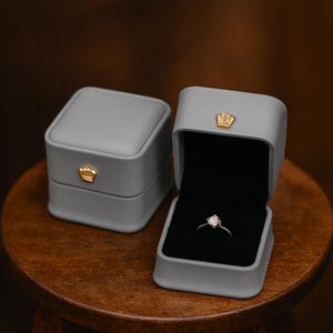 May include: Two gray ring boxes with gold crown accents. One box is closed, the other is open, revealing a diamond ring set against a black interior. The boxes sit on a wooden surface, suggesting a jewelry display.