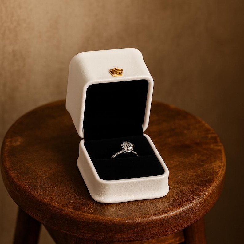 May include: A white ring box with a gold crown emblem on top, open to reveal a diamond ring on a silver band. The box has a black velvet interior. The box is sitting on a wooden stool.