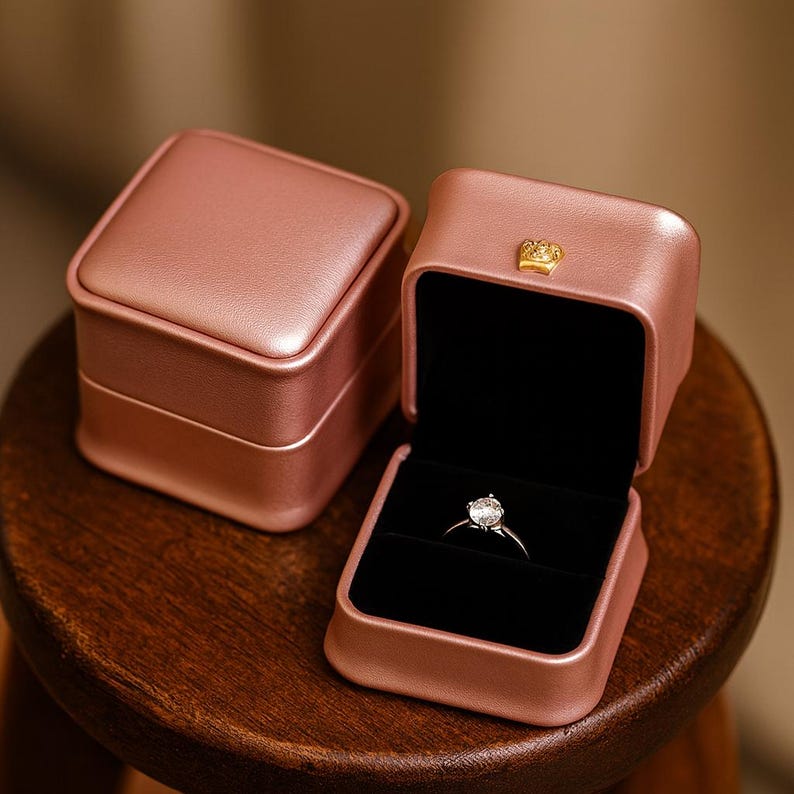 May include: A rose-colored, square ring box with a gold crown detail, open to reveal a diamond ring on a black velvet interior. Another closed box is visible. The boxes are on a wooden surface.