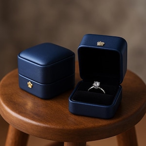 May include: Two navy blue ring boxes, one closed and one open, displaying a diamond ring. The boxes have a gold crown emblem. The open box reveals a black velvet interior, showcasing the ring. The boxes sit on a wooden stool.