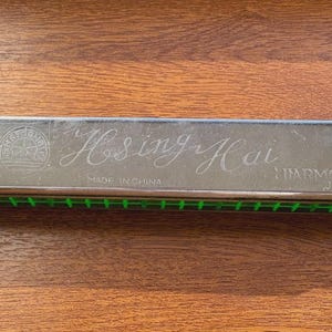 May include: A silver Hsing Hai harmonica with green accents, on a wooden surface. The harmonica has the words "Hsing Hai Harmonica" engraved on its surface, along with "Made in China".