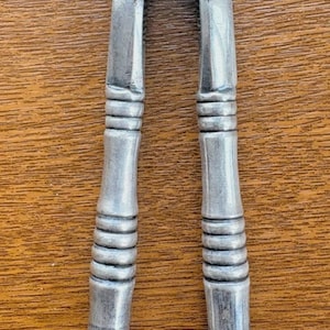 May include: A silver-coloured metal nutcracker featuring two long, parallel handles with a series of horizontal ridges and grooves along the length. The top section includes two circular pivot points connected by a central hinge mechanism for cracking nuts.