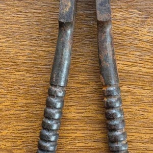 May include: A pair of vintage metal nutcrackers featuring a dark, oxidised finish with visible rust spots. The tool has two long, tapered handles with a ribbed, textured grip section and a hinged head designed for cracking shells.