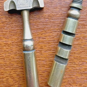 May include: A two-piece brass tool featuring a T-shaped head with an engraved profile of a person, the text "TRADEMARK" and "VERY BEST," and a cylindrical handle. The second piece is a matching brass cylindrical rod with a threaded top end.