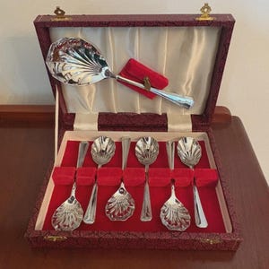 May include: A collection of silver serving spoons and a large serving spoon, displayed in a burgundy box with a cream satin lining. The spoons feature a shell design and are presented on a red velvet base.