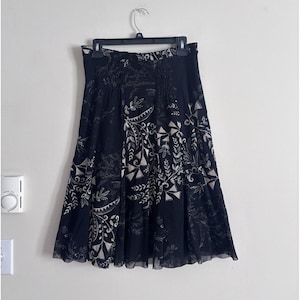 May include: Black skirt with a white floral and geometric pattern. The skirt is hanging on a black hanger. The skirt has a gathered waist and a flowing, flared shape. The fabric appears to be lightweight and semi-sheer.
