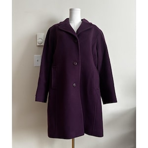 May include: A deep purple, knee-length coat featuring a notched collar, long sleeves, and a front button closure with three matching dark buttons. The garment has two vertical side pockets and is displayed on a white mannequin torso.