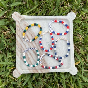 May include: Five colorful beaded bracelets with team names, including "EAGLES", "JAGS", "BULLS", "DALLAS", and "SAINTS", are displayed on a marble tray. The bracelets feature various color combinations, such as green and white, red and blue, and yellow and black.