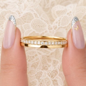 Könnte beinhalten: Ein goldener Ring mit einer dünnen Schiene, in die auf der oberen Hälfte eine Reihe kleiner, klarer, rund geschliffener Steine eingelassen ist. Das Metall hat eine polierte, reflektierende Oberfläche und ein schlichtes, schmales Design.