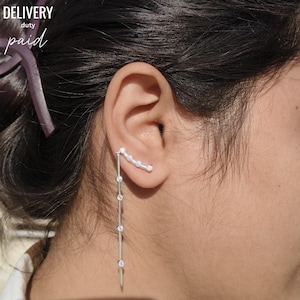 May include: A silver ear cuff and dangling earring set. The ear cuff curves along the upper ear with small, clear stones. The earring dangles down with a series of clear stones. The words "DELIVERY duty paid" are in the upper left corner.