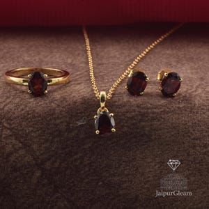 May include: A gold-coloured jewellery set comprising a ring, pendant necklace, and stud earrings. Each item displays a dark red oval gemstone. The ring and earrings have a simple design, while the pendant hangs from a fine chain.