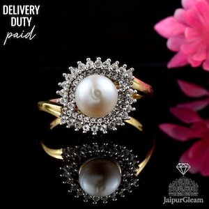 May include: A gold ring with a large, round, white pearl at the centre, encircled by a halo of smaller, clear gemstones. The ring's band is gold, set against a black backdrop. The text "DELIVERY DUTY paid" is visible.