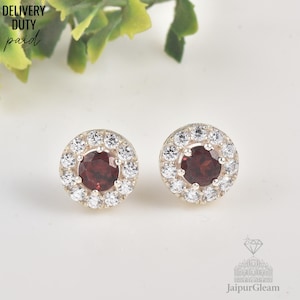 May include: A pair of round stud earrings. Each earring has a central, deep red gemstone encircled by a halo of smaller, clear gemstones. The earrings are set in a silver-coloured metal. The text "DELIVERY DUTY paid" is visible.