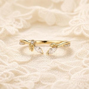 May include: A gold open ring featuring a delicate branch design with small leaves and clear marquise-cut stones. The band is accented with tiny round stones along the sides, creating a symmetrical botanical appearance with an open centre gap.