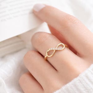 Puede incluir: Un anillo de tono dorado con un diseño de símbolo de infinito. El centro del bucle de infinito está adornado con una fila de pequeñas piedras claras y brillantes. La banda es delgada y pulida, creando un acabado metálico suave y continuo.