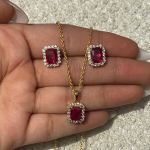 Puede incluir: Un conjunto de joyas de color dorado que incluye un collar y pendientes a juego. Cada pieza tiene una piedra preciosa rectangular de color rojo intenso rodeada de pequeñas piedras transparentes. El colgante del collar y los pendientes están engarzados en metal dorado.